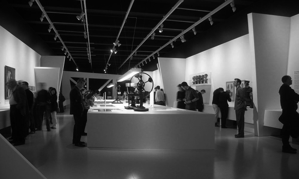 Menschen betrachten Kunstwerke in einem Museum, umgeben von verschiedenen Ausstellungen und informativen Beschilderungen.