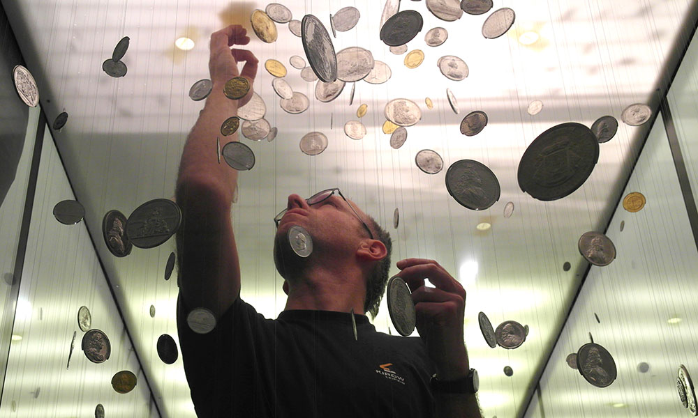 Eine Person steht unter hängenden Münzen unterschiedlicher Größe und Farbe und streckt eine Hand nach oben. Die Münzen hängen an dünnen Drähten in einer gut beleuchteten Vitrine.