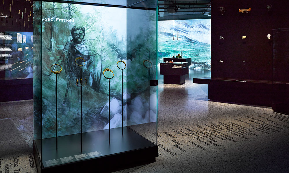 In einer Glasvitrine in einem Museum werden antike Schmuckstücke ausgestellt. Hinter der Vitrine ist ein gemalter Hintergrund mit einem Mann in historischer Kleidung zu sehen, an den Wänden hängen weitere Artefakte.