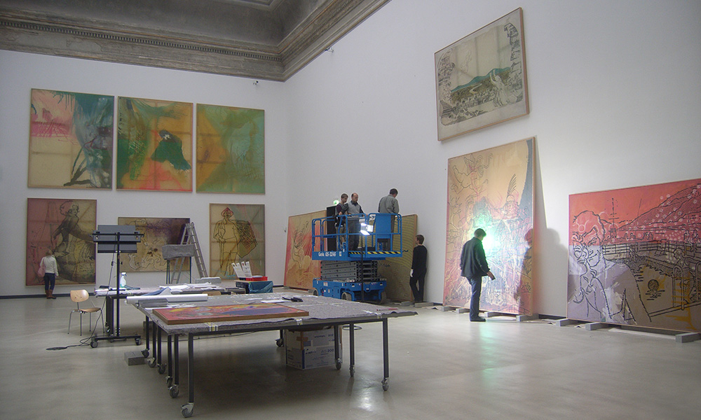 Ein Mann arbeitet an einem Gemälde in einem hellen Raum mit Kunstmaterialien um ihn herum.